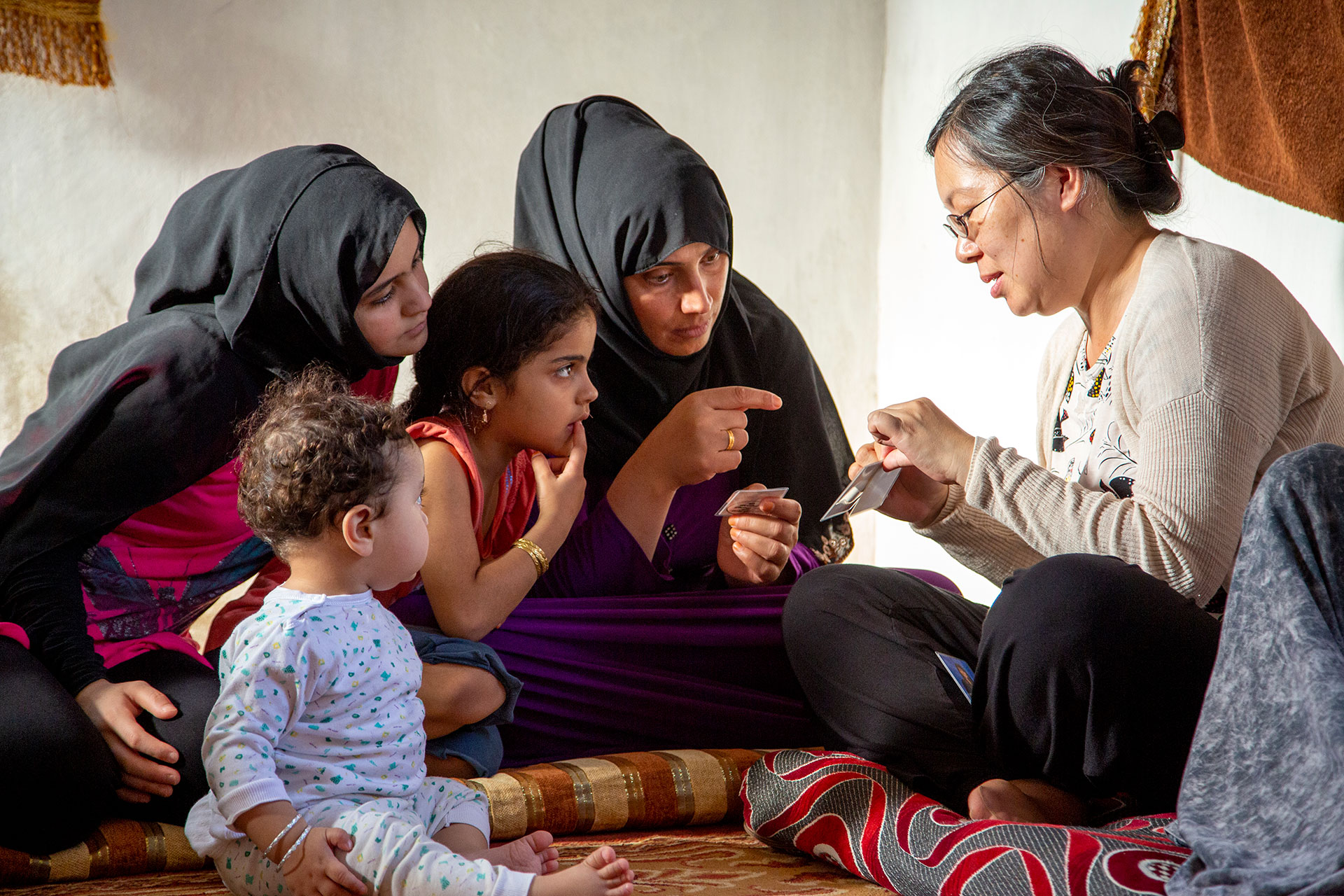 Asian American female missionary telling a story, sitting on cushions with children and mothers wearing hijabs